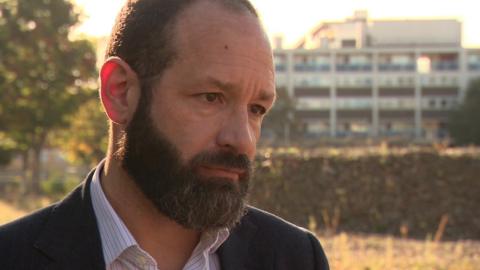 Southwark Council leader Kieron Williams in a striped shirt and dark suit jacket. He has short dark hair, a moustache and beard. He stands in front of an out of focus building. 