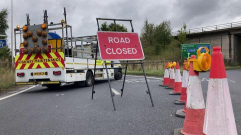 A red road closed sign sits behind a row of roadworks which is blocking off a slip road to the motorway. A highway maintenance van is also visible. 