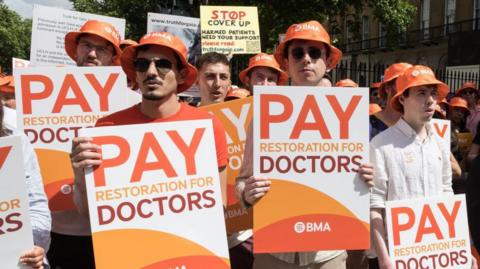 A group of people in orange bucket hats hold "pay restoration for doctors" signs