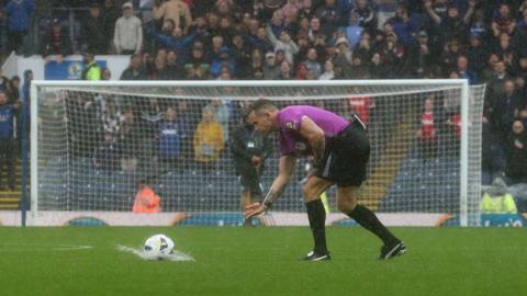 Referee wearing a purple t shirt, black shorts and black socks stands on the green football pitch bent over looking at the ball on the ground which he rolls as it makes a splash.