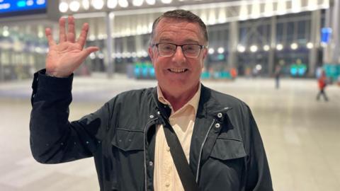 Kieran Doherty stands in Grand Central Station, smiling and waving at the camera. He is wearing glasses, a cream shirt and a black jacket. The background of the photo is blurred. 