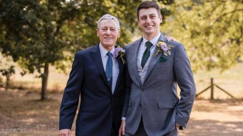 Robert on the left wearing a dark suit with blue checkered shirt and tie with a flower pinned to it and Simon with his arm around his shoulder also wearing a dark suit with a flower pinned to it. They are both smiling at the camera and stood in front of trees.