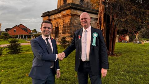 Two men wearing dark suits are seen shaking hands. They are stood in what appears to be a churchyard.