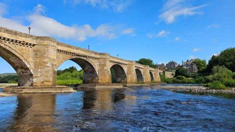 A viaduct over a river with trees and buildings in background under a blue sky