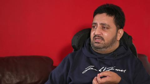 Ahsan Razzaq, wearing a dark blue hooded top, is sitting in his wheelchair in the living room of his home in Oldham. 