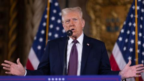 Donald Trump speaking at rostrum. He is wearing a blue suit and reddish-coloured tie and parting his hands. Two US flags are in the background