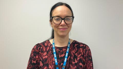 A smiling Fay Wickett looks directly at the camera, she is wearing a red-patterned top with a blue NHS lanyard around her neck. She has long brown hair tied back and black-rimmed glasses.  