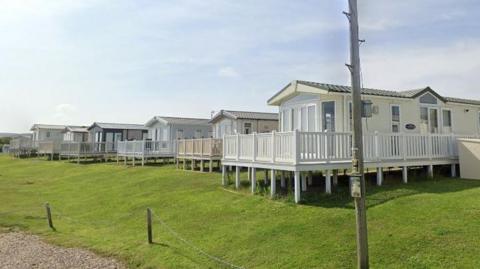 A row of modern static caravans raised on stilts along a grassy slope, each with a white‑railed deck, a gravel path and rope fence in the foreground, and a utility pole standing nearby.
