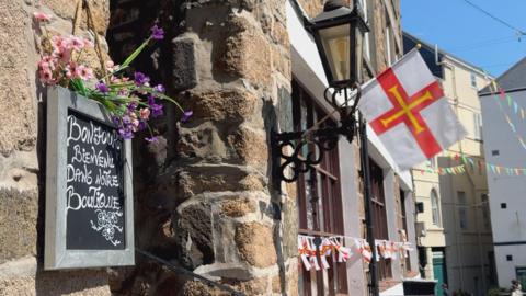 A chalkboard sign reading “Bonjour, Bienvenue dans notre Boutique” mounted on a stone wall beneath pink and purple flowers. A Guernsey flag hangs from a black lantern bracket, with bunting visible along the street in the background