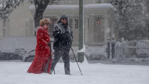 A man and woman in thick winter coats walk through a snow flurry with walking sticks