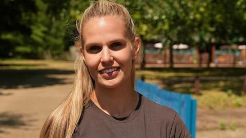 Kirsty Gillingham smiles for the camera. She is standing outside on a sunny day. She has long fair hair and is wearing a brown top.