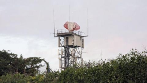 A metal tower is seen rising above a bush in the foreground. The tower has a red and white radar dish on the top.