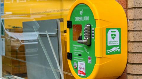A bright yellow public defibrillator cabinet is mounted on a rough grey brick wall. The box has a small window, a keypad for entry, and a black handle. Text on the front reads “Life Saving Defibrillator” and instructs people to call 999 for an access code. A green sign with a heart and lightning bolt symbol indicates the presence of a defibrillator, and a St John Scotland logo appears at the bottom.