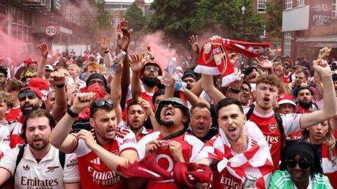 A large group of Arsenal fans celebrate - red flare smoke is in the background, while lots of people in red and white Arsenal shirts and flags celebrate