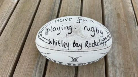 A rugby ball lying on wooden decking. The ball is a white Gilbert ball and appears well worn. A message has been written on in black market pen. It reads, Have fun playing rugby - Whitley Bay Rockcliff.