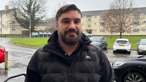 A man in his 40s with short brown hair swept to the side and dark facial hair smiles as he stands in the rain on a housing estate. Behind him is a road with cars parked on it, a large grass verge and a three-bed block of light coloured flats.