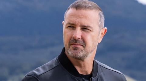 Paddy McGuinness, with short dark hair and a stubbly beard, wears a black zip-up leather jacket and stands in front of a tree-covered hill