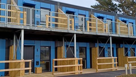Exterior of the modular units in Cornwall. The terraced units are blue and black. They have wooden fences in front of their entrance.