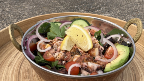This picture shows a metal bowl of salad placed on a wooden tray with a beach in the background. The salad includes sliced avocado, cherry tomatoes, red onion rings, leafy greens, grilled or cooked octopus pieces and two lemon wedges.