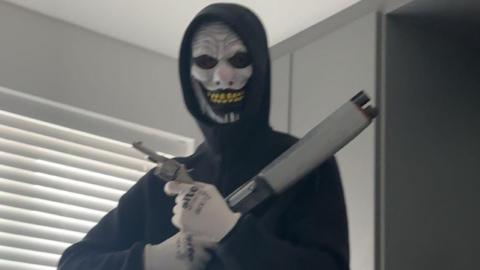 A masked man wearing a black hooded top and white gloves while holding a shotgun and a handgun. He is standing indoors in front of a window which is covered with blinds.