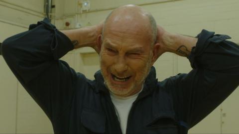 A still from the upcoming film, All You Do Is Bleed. Ian Hodson, a bald man with a short beard, is screwing his face up in distress with his hands behind his head.