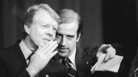 Jimmy Carter, with a hand over his mouth as if to speak in a noisy room, leans towards Joe Biden who is pointing into the distance. Both men are wearing suits and appear to be on stage. The photo was taken at a fundraiser in Wilmington in 1978