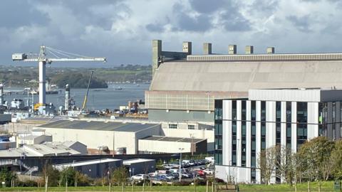A general view of of the dockyard. Large buildings and a crane are visible. Water and boats are in the distance. The sky is grey and cloudy.