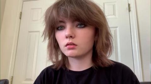 Rebecca Young has brown hair, grey blue eyes and is wearing a black t-shirt. She is sitting in front of a white door and is chatting over Zoom.