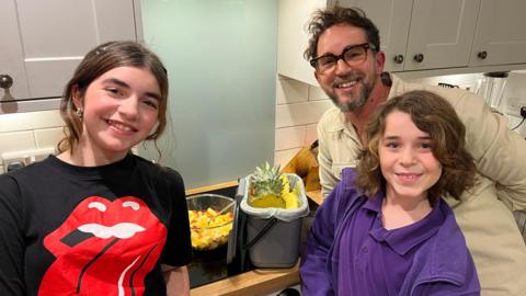 A father and two children stand smiling in their kitchen beside a bowl of fruit salad and their kitchen caddy filled with fruit peelings.