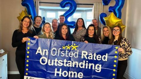 Seven women and four men are picture in a room in a residential property holding helium filled balloons with the numbers 1, 2 and 3 and displaying a large banner which says "An Ofsted rated Outstanding Home"