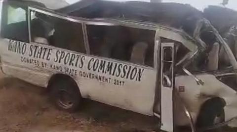 A badly damaged Kano State Sports Commission bus, with a crumpled front end and roof partially torn off