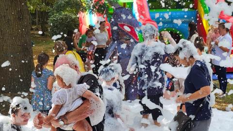 Children and parents playing with white foam- with a colourful bouncy castle in the background.