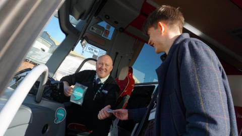 A male bus driver takes a fare from a male passenger. The driver has a sticker in his hand showing that he speaks Gaelic. Both driver and passenger are smiling.
