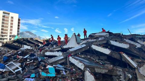 A group of about ten rescuers in orange uniforms and helmets are working on top of a pile of rubble with large concrete slabs and smaller items. Behind, a block of flats remains standing, and a mountain is visible in the background, set against a blue sky with light white cloud.
