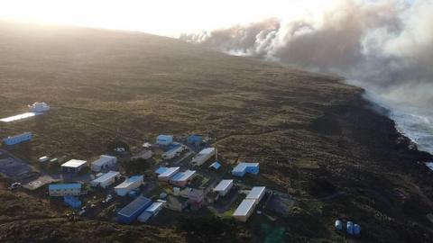 A coastline with large plumes of smoke in the distance. In the foreground there is a small collection of mobile buildings making up the La Roche Gordon research station. 