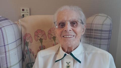 A woman sitting in a tartan armchair, smiling at the camera. She is wearing a white, collared blouse and clear-framed glasses. She has fine grey hair tied back.