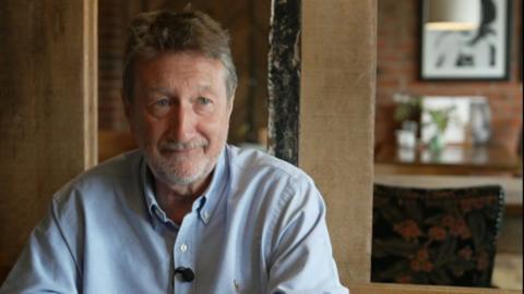 Steven Knight, wearing a blue shirt, is sat in room with a wooden pillar and behind is a chair with ornately-patterned fabric. In the distance are images attached to a bare-brick wall.