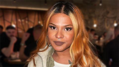 A close up photo of Seana Momsen in a restaurant with people sat at tables visible in the blurred background. She has bronze and blonde coloured hair and has a diamond nose ring in and a hoop earring in her right ear.