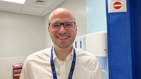 A smiling man with a bald head and glasses in a white shirt with a blue lanyard in a room with white walls and a blue curtain