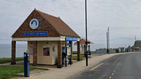 A small, cliff-top brick building with a blue and white sign reading "Lift to West Cliff Beach".