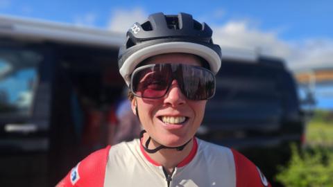 Laura is wearing a black helmet and cycling goggles and is smiling at the camera. She is wearing a white and red cycle jersey. 