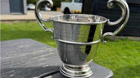 The silver trophy is engraved with Grimsby and District Cricket League and has two substantial handles. It is sitting on a table at the club, reflecting the green grass on a sunny day.