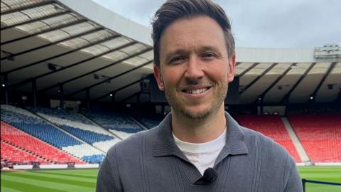 Lewis Irons stands by the pitch in Hampden stadium, he wears a white tee-shirt and a grey top