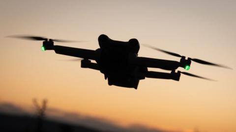 Silhouette of a small drone featuring four small propellers, set against a darkening sky. Two green lights are visible on the drone.