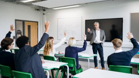 A classroom full of school children in a lesson with a teacher at the front of the class