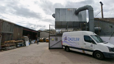 The entrance and yard of a construction company. A van is in the foreground with the name Oastler on the side. Pallets and wood are in the yard