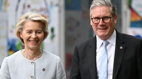 A man and woman look towards the camera. The man is wearing a dark suit, white shirt and light blue tie. He has grey hair and is wearing dark rimmed glasses. The woman has short blonde hair and is wearing a light grey buttoned jumper. Both are wearing badges in the shape of a small star.