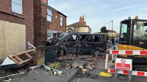 A house is partially boarded up and there are bricks strewn across the road. A digger with a sign reading 'no smoking' is also in the middle of the road