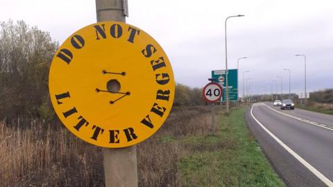A yellow circle-shaped metal sign with ' do not toss litter' written on it in black, placed on a lamppost on the side of the road.