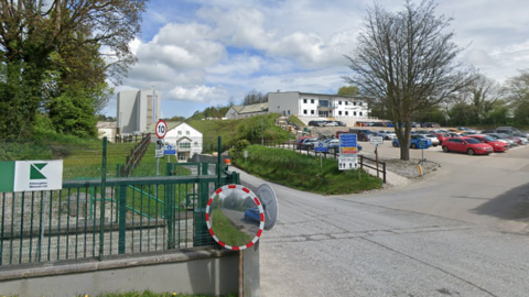 The Kilwaughter Minerals' building is white and is pictured from the road side. The green metal fence with the company's logo is in shot and so is a car park with multiple cars in it.